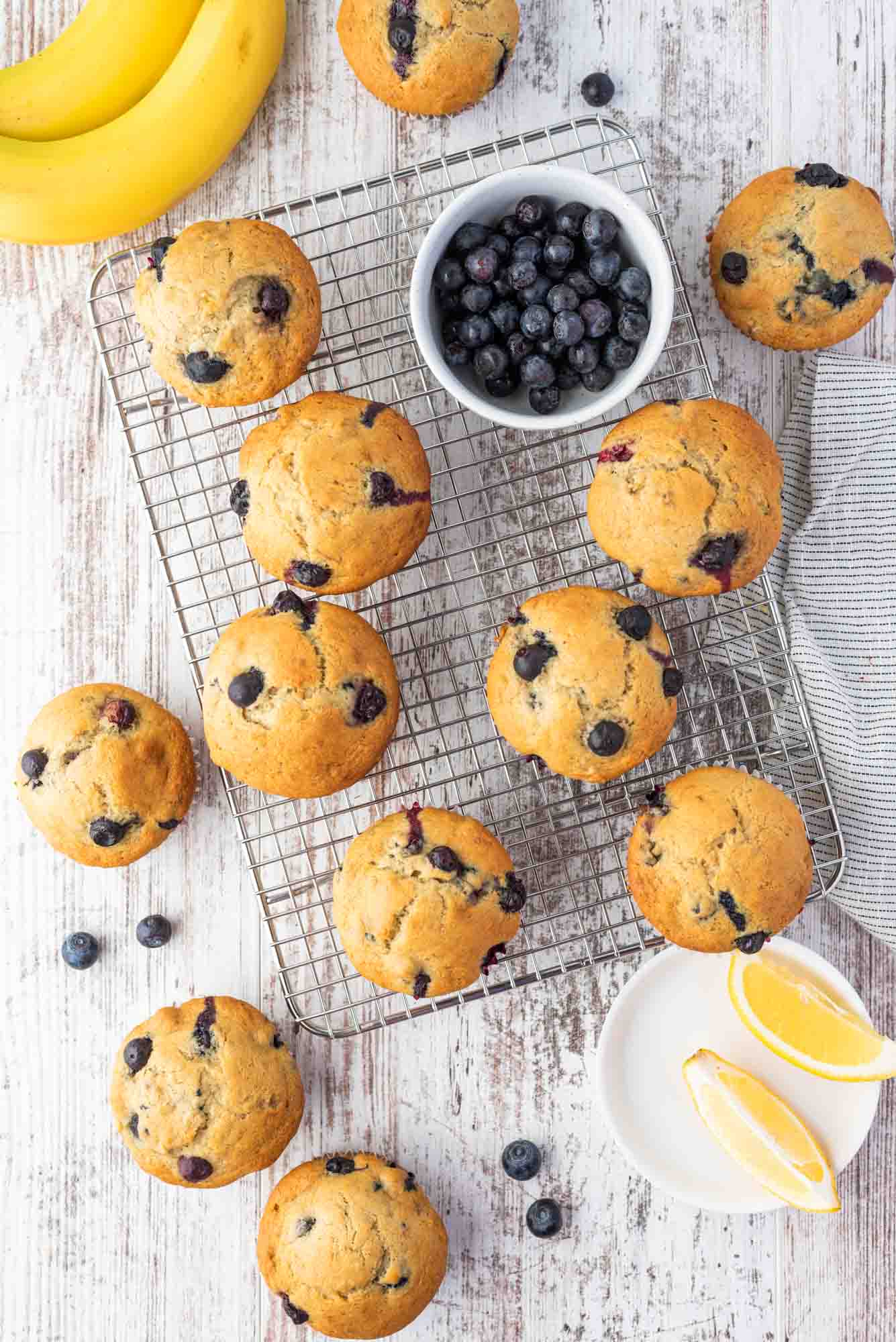 Overhead shot of muffins on a wire rack, with fresh blueberries, lemon wedges, and bananas on the side.