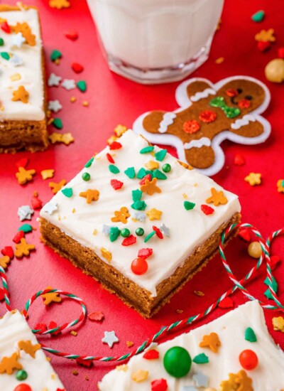 Frosted gingerbread cookie bars with holiday sprinkles on a red table. A glass of milk is in the background