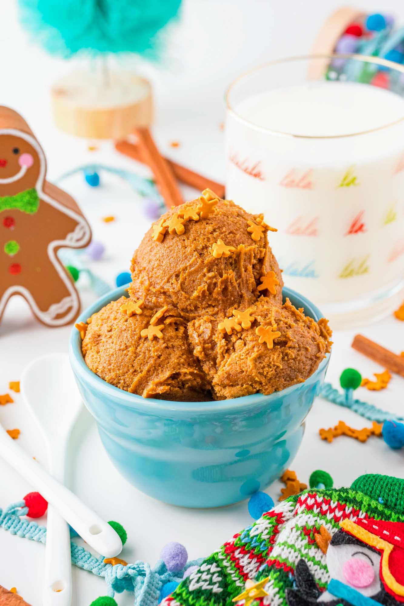 a blue bowl with three scoops of gingerbread cookie dough topped with gingerbread man sprinkles. The bowl is surrounded by christmas themed decorations. 