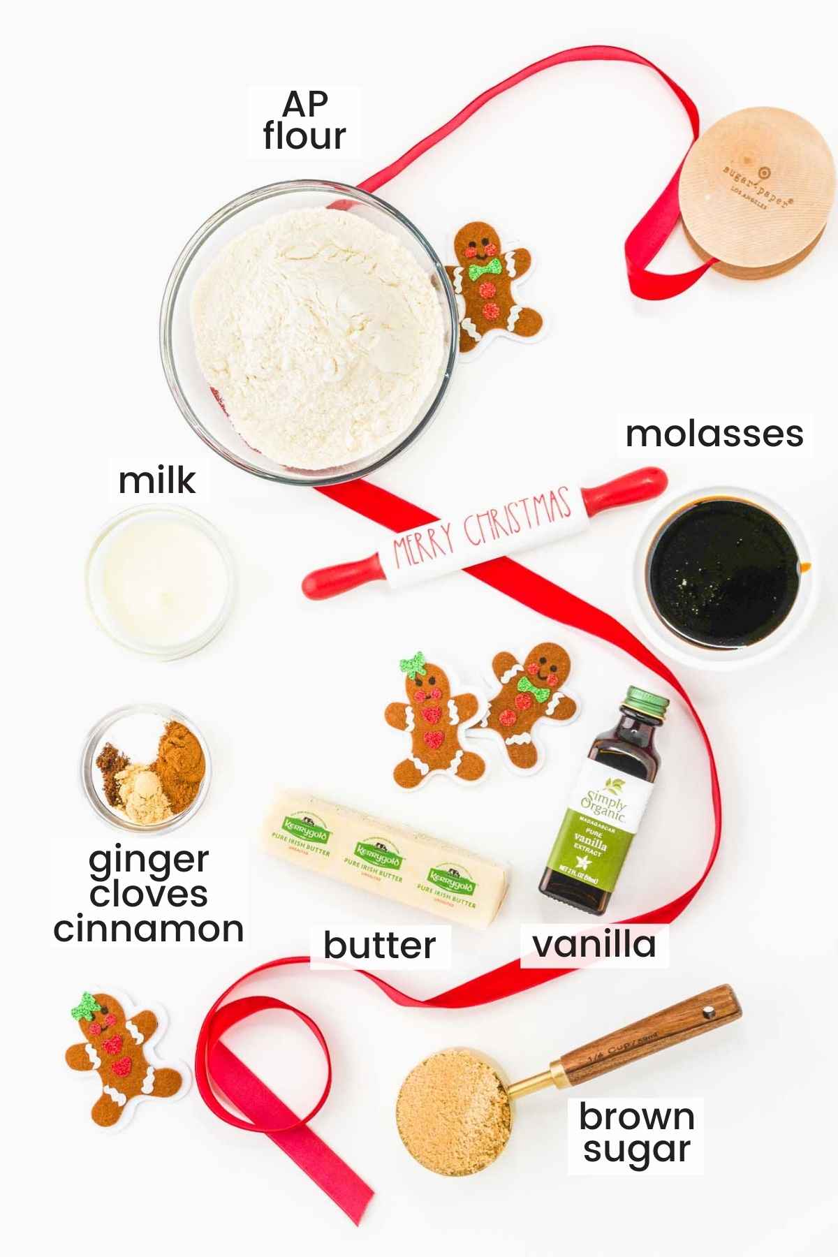 the ingredients for gingerbread cookie dough on a white counter in separate bowls. A red ribbon and gingerbread men decorate the counter.