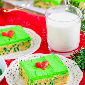 Two plates of grinch cookie bars on white plates sit on a red clothed table next to a glass of milk and a string of fairy lights.