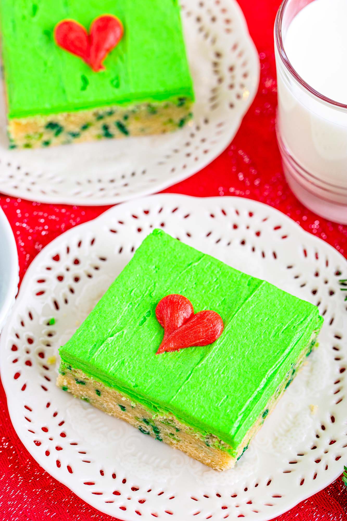 two cookie bars with green grinch icing and red hearts drawn in icing on paper doilies on a red table.