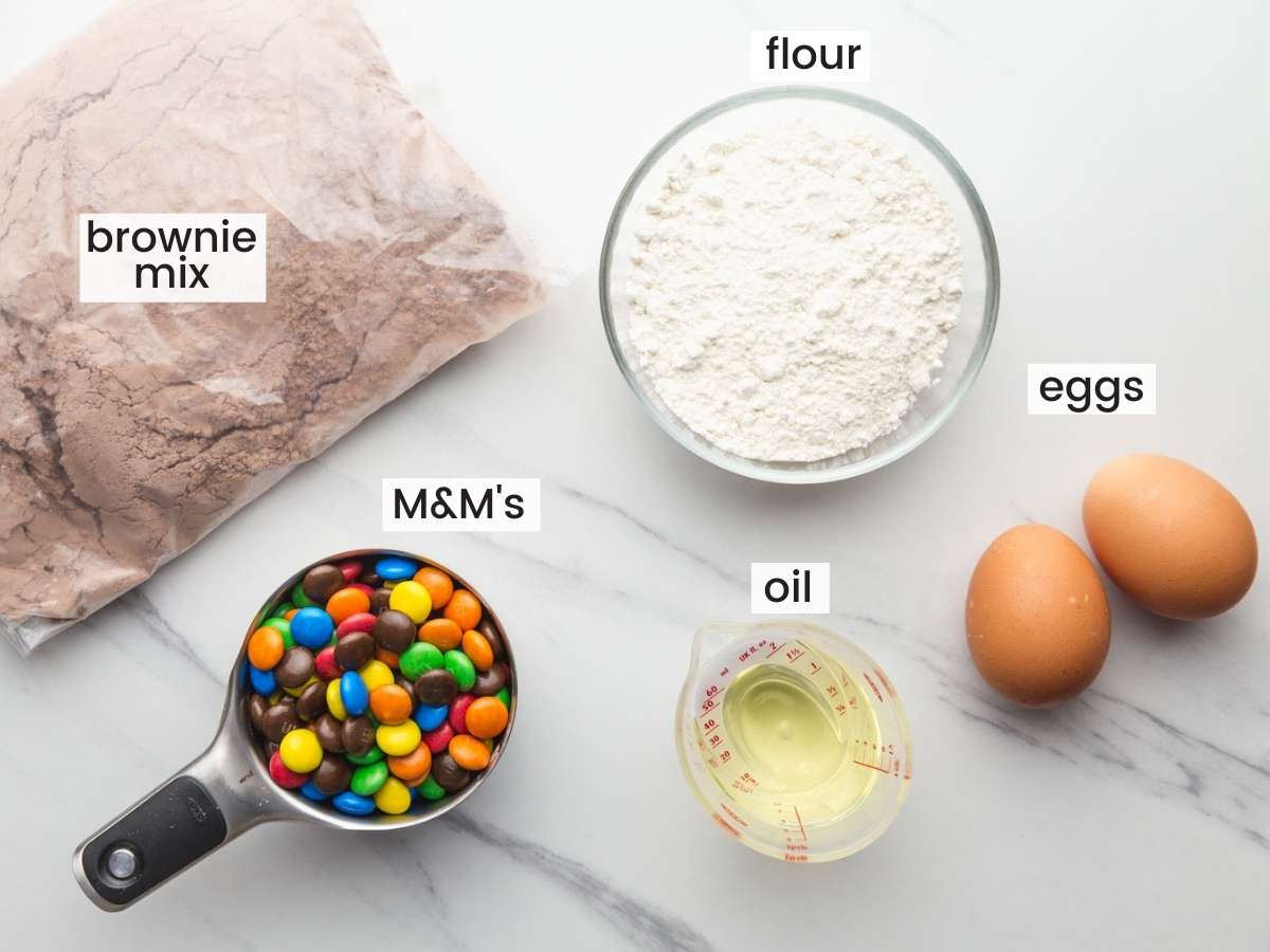 the ingredients needed to make brownie cookies, including a boxed brownie mix, m&ms candy, flour, oil, and eggs
