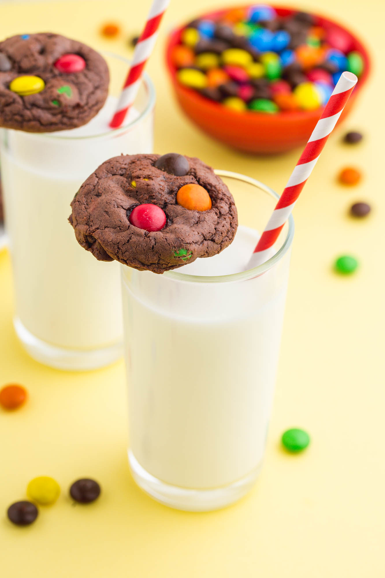two glasses of milk with red and white striped paper strays and chocolate m&m brownie cookies balanced on top.