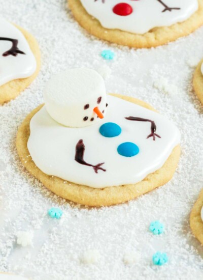 a round sugar cookie decorated with a marshmallow to look like a melting snowman. The cookie is sitting on a plate of sugar that looks like snow.