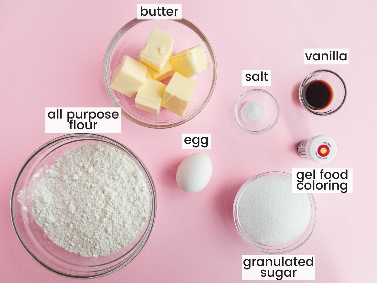 Ingredients for spritz cookies in separate bowls on a pink table.