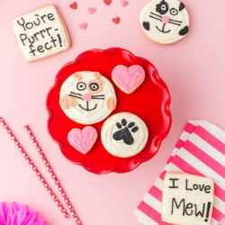 A pink table with a red cake riser topped with round cookies decorated like cats and pink heart cookies. Square cookies to the side say "you're Purrfect" and I Love Meow!