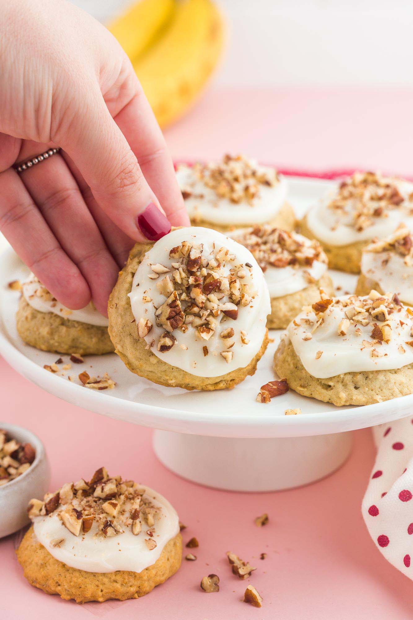 a cake stand filled with banana bread cookies with white frosting and chopped nuts. a feminine hand is picking a cookie up.