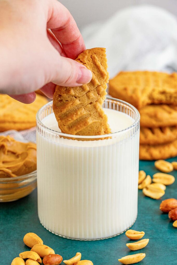 a short glass of milk. A hand is dipping half of a peanut butter cookie into the glass. In the background there are stacks of cookies and shelled peanuts.