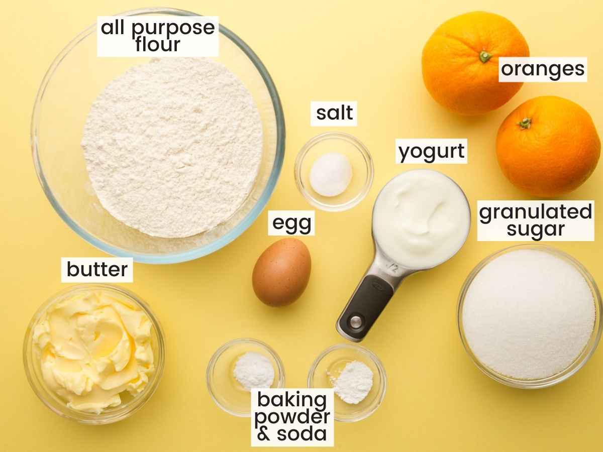 The ingredients for making Orange Cookies measured into separate glass bowls on a yellow background. Flour, oranges, sugar, yogurt, butter, egg, and others