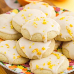 a bright floral plate filled with soft orange cookies topped with glaze and orange zest. In the background is halved oranges