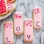 four tall rectangle cookies decorated with pink royal icing and sprinkles. Letters spelling LOVE are written on the cookies, one letter on each.
