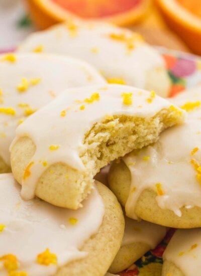 a closeup view of a platter of soft orange cookies with icing, the one on the top has a bite taken. Orange halves are in the background.
