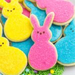 easter bunny cookies decorated with pink, blue, and yellow icing and sugar with peeps bunny faces, on a platter lined with green easter sprinkles.
