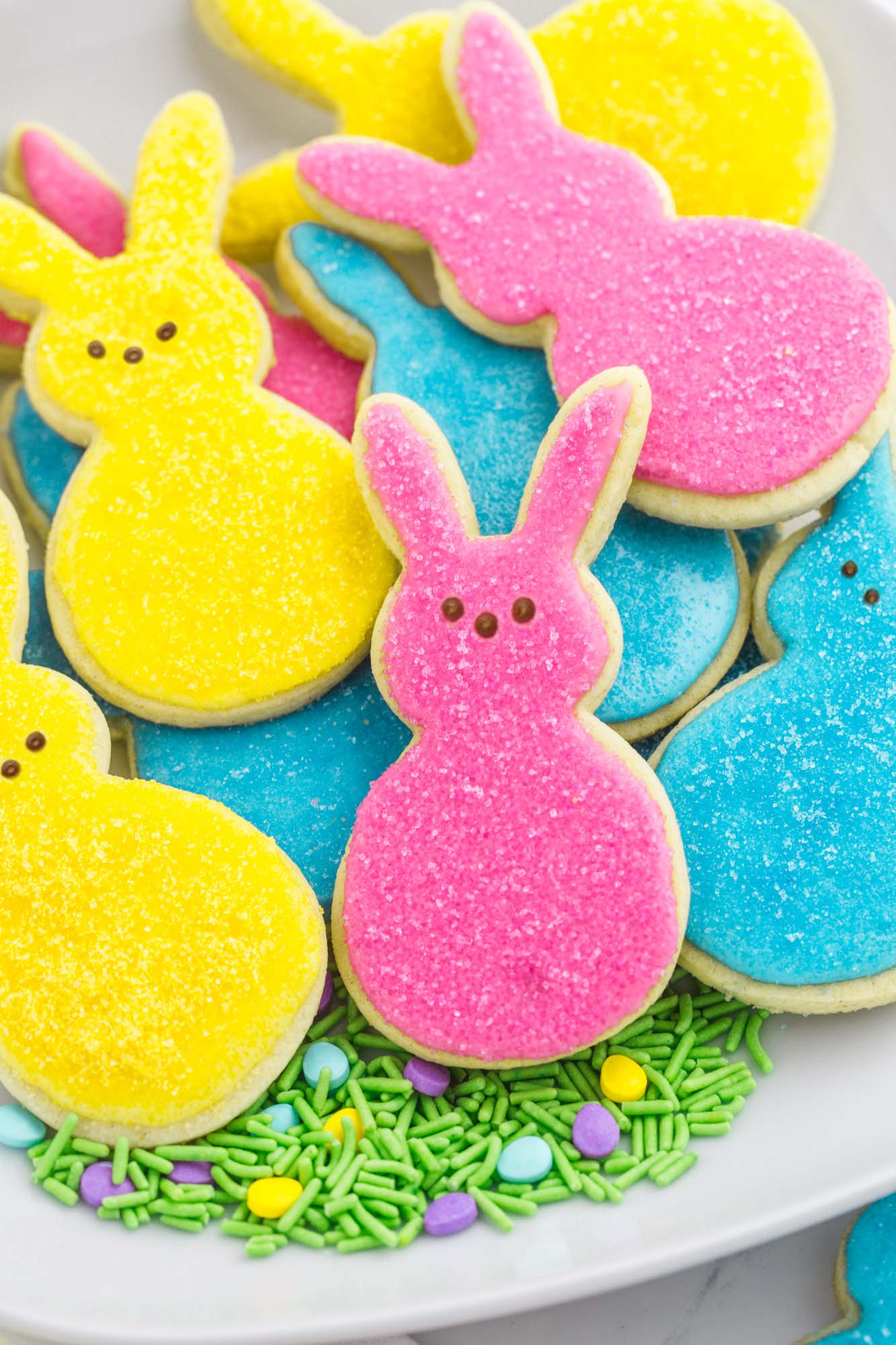 easter bunny cookies decorated with pink, blue, and yellow icing and sugar with peeps bunny faces, on a platter lined with green easter sprinkles.