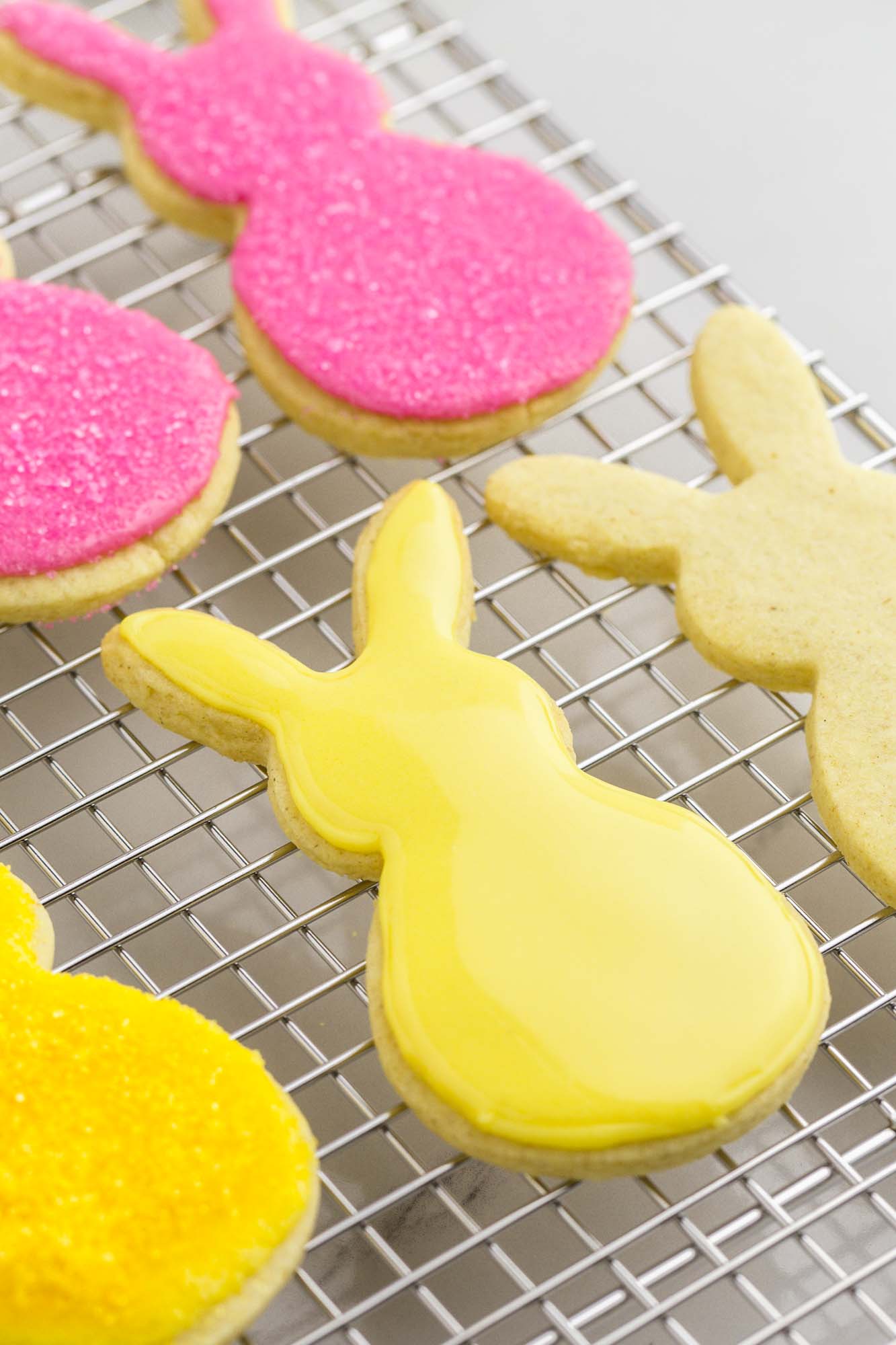 a row of yellow iced easter bunny cookies and a row of pink iced easter bunny cookies on a wire rack