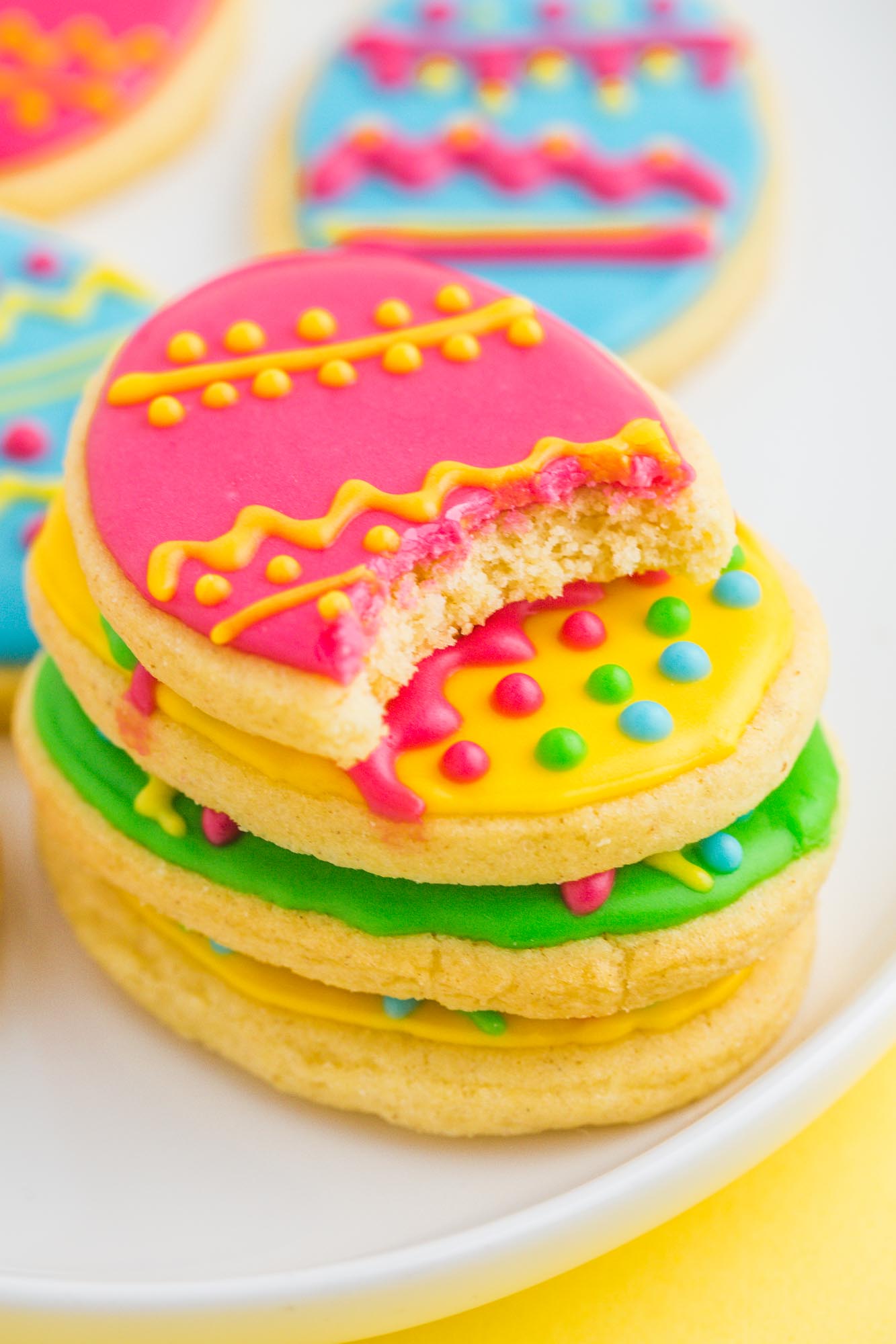Four easter egg cookies stacked on top of one another. The top cookie, a bright pink one with yellow details has been bitten into. 