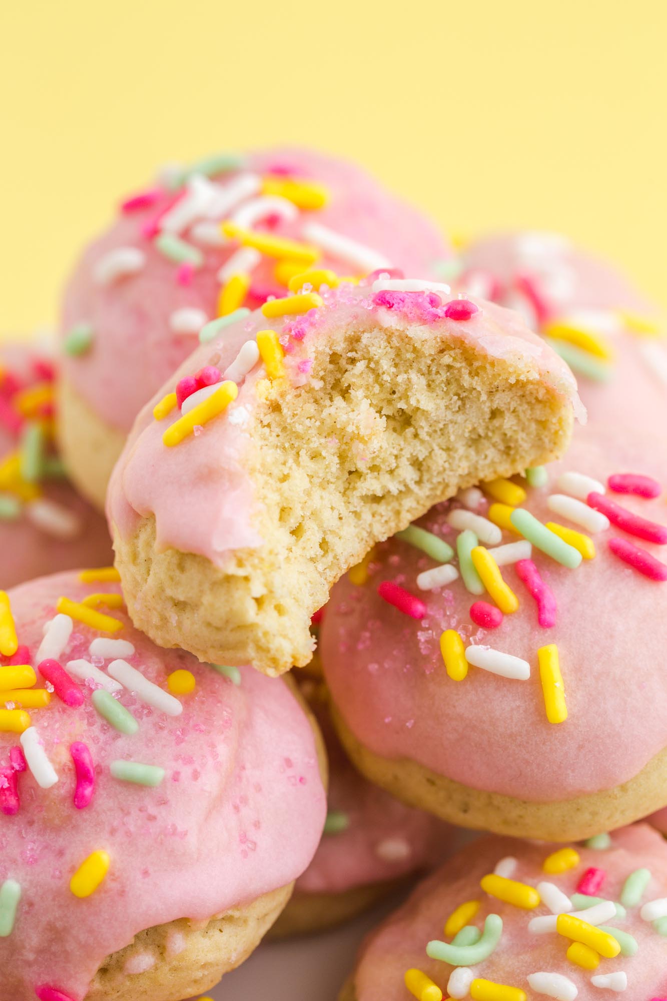 closeup view of a pile of italian cookies with pink frosting and sprinkles. One has had a bite taken, showing the texture inside.