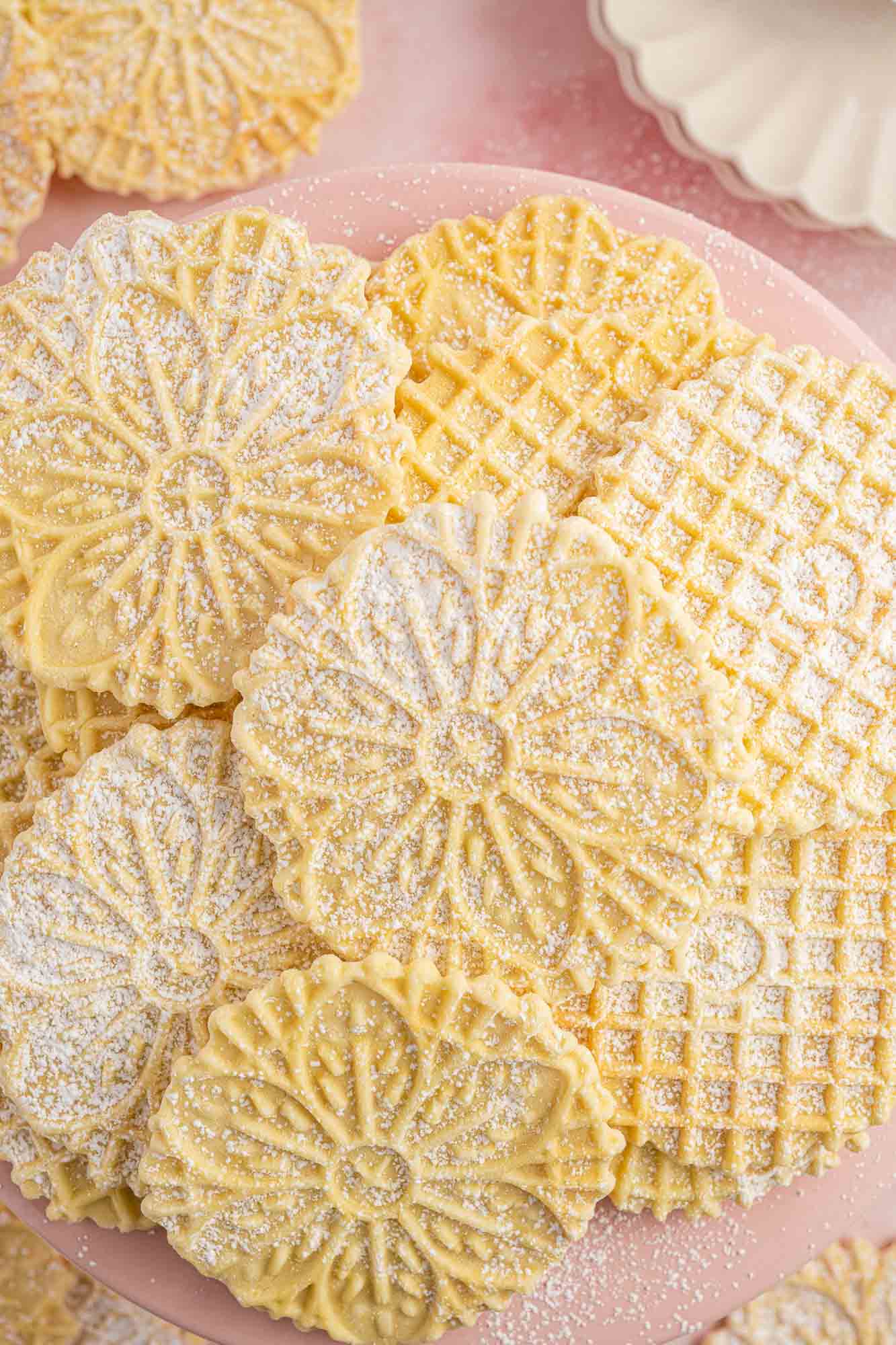 a pile of thin pizzelle cookies on a plate, dusted with powdered sugar