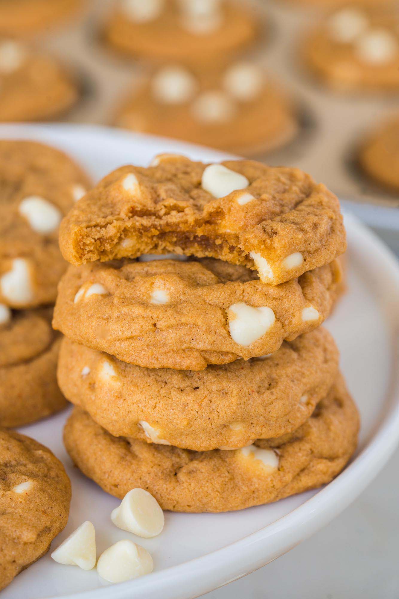 a stack of four pumpkin spice cookies on a plate, one has a bite taken out. White chocolate chips are next to the cookies