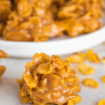 A rounded peanut butter cornflake cookie on the counter in front of a plate of cornflake cookies.