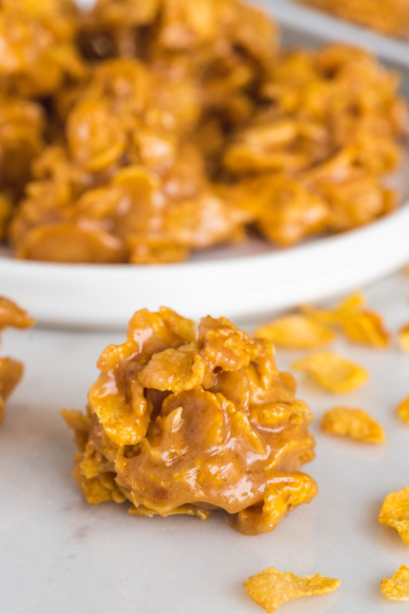A rounded peanut butter cornflake cookie on the counter in front of a plate of cornflake cookies. 