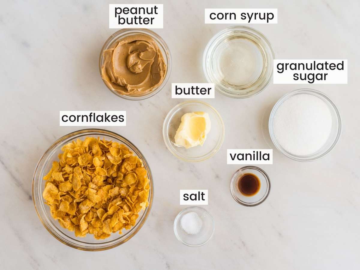 The six ingredients in cornflake cookies, in separate glass bowls on a marble counter, viewed from above.