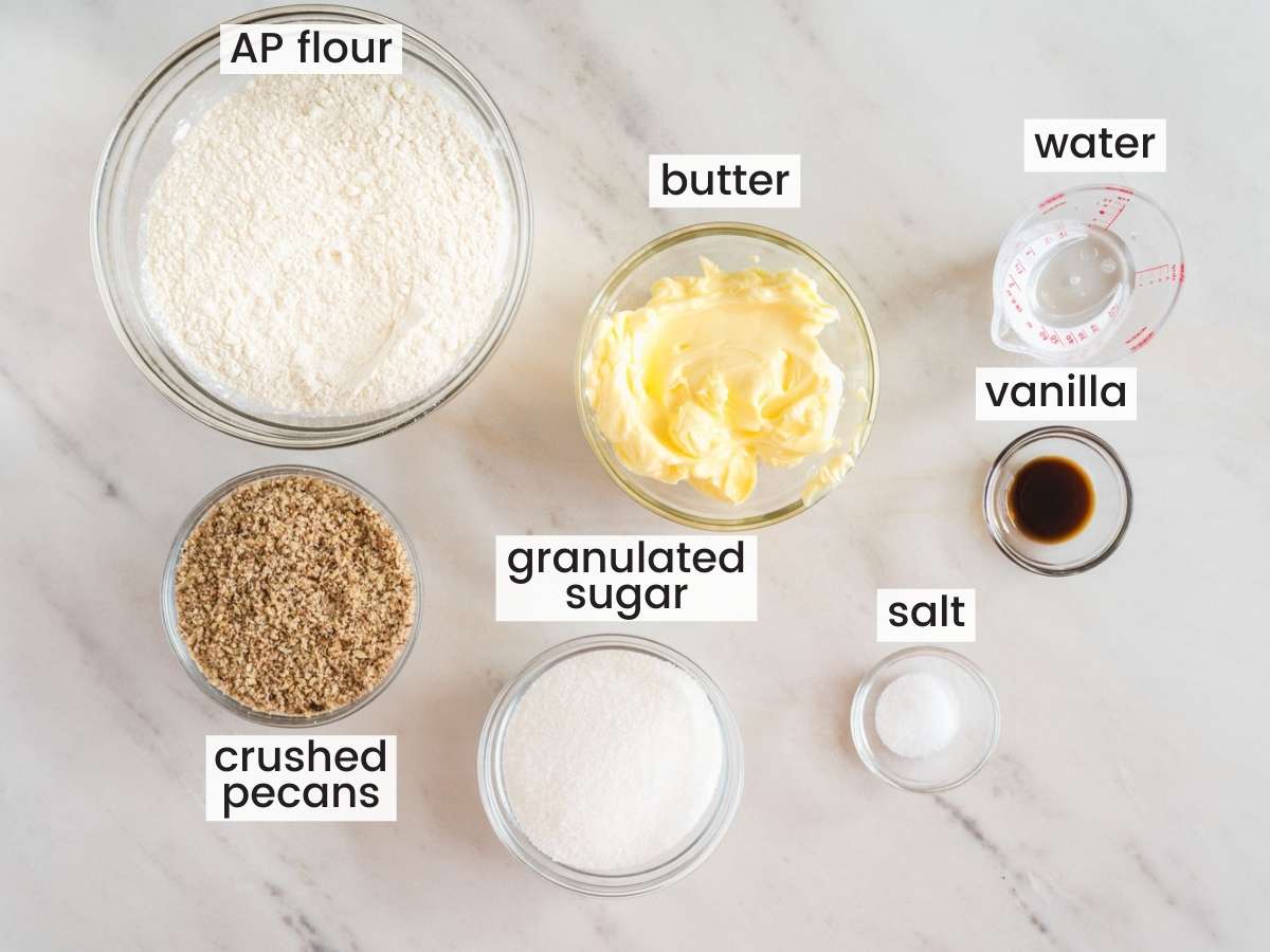 The ingredients for homemade pecan sandies cookies, measured into separate bowls on a marble counter. Viewed from above