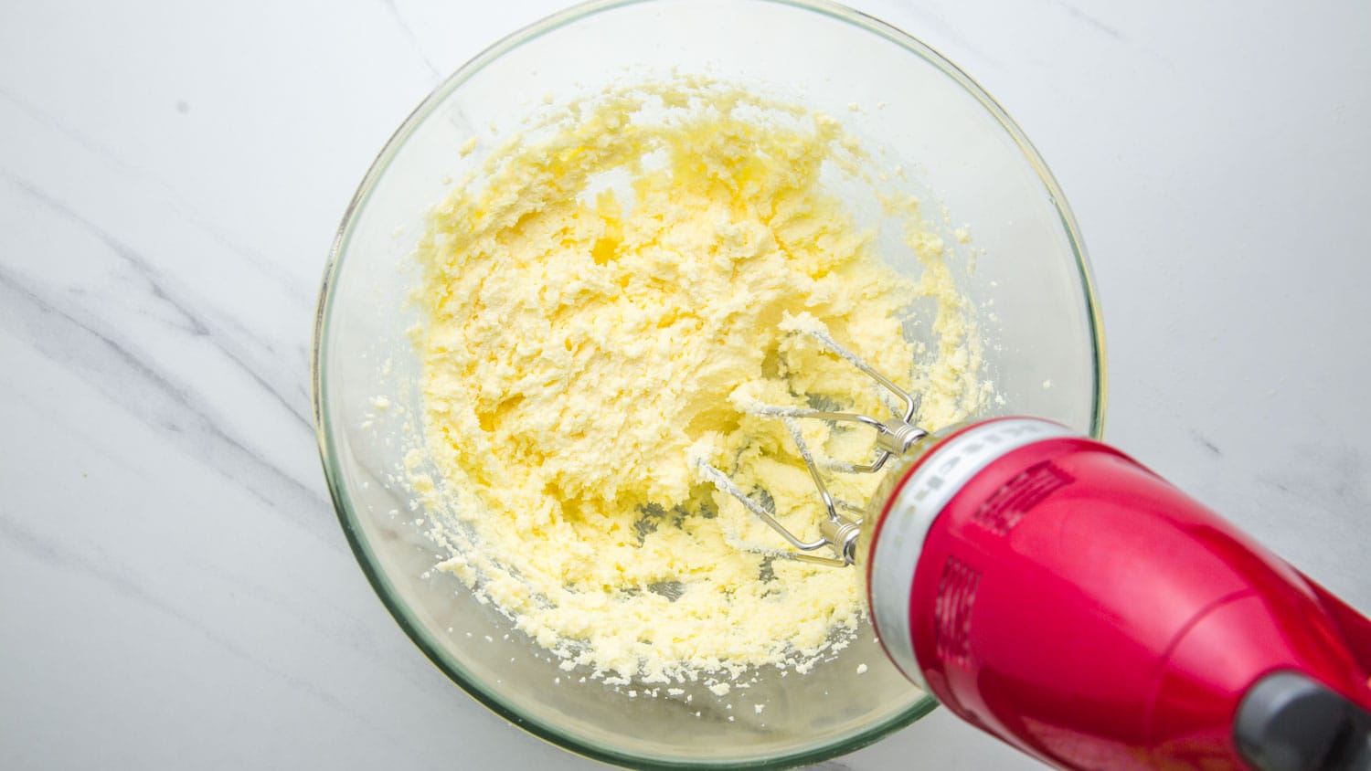 Creamed butter and sugar in a glass bowl, and a red kitchenaid hand mixer.