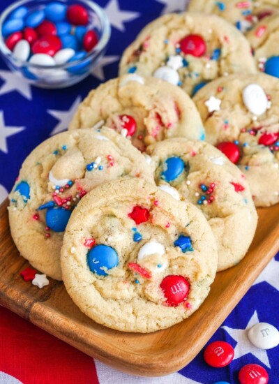 Red white and blue M&M cookies placed on a wooden plate, with USA flag in the background.