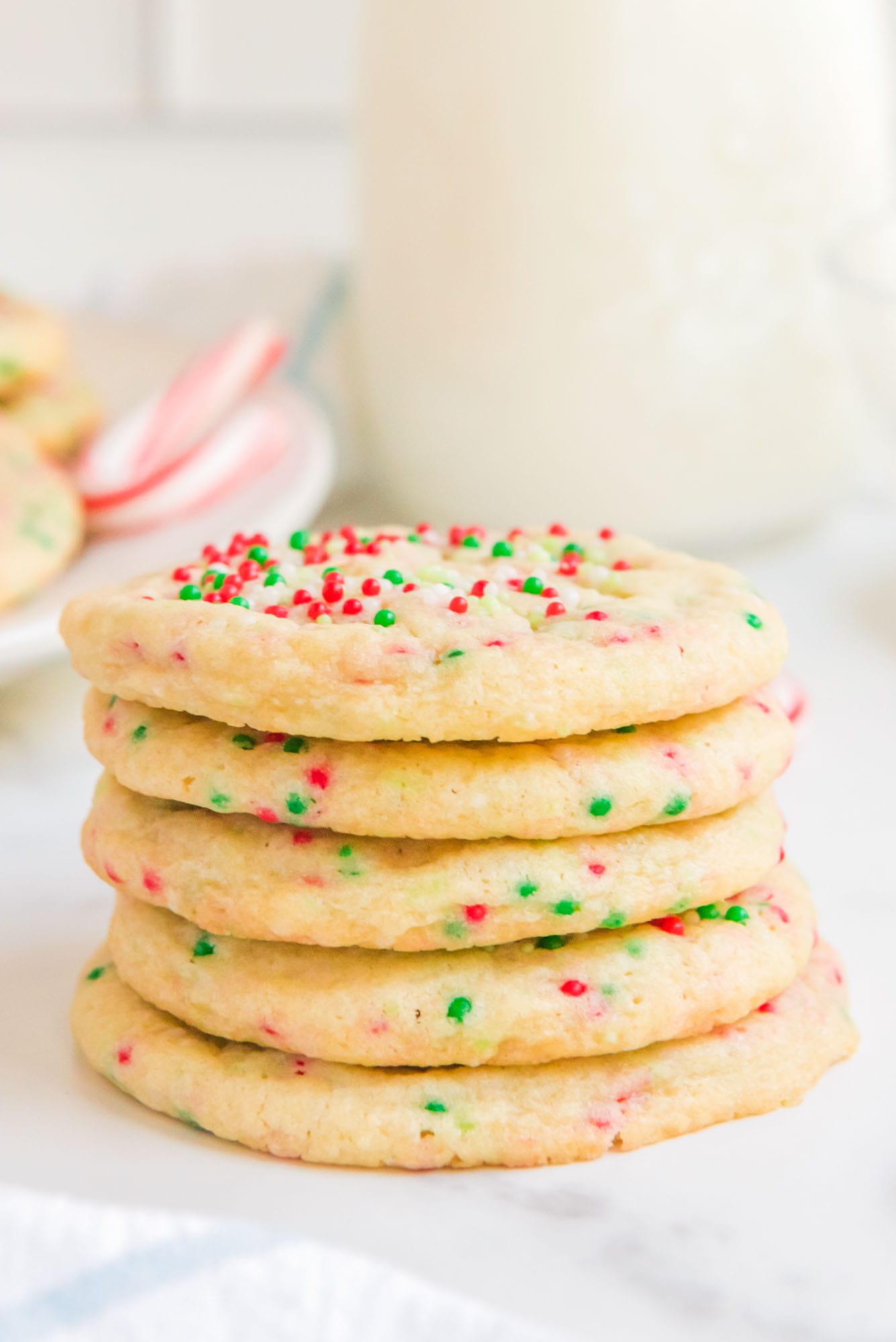Stacked sprinkle cookies on a white counter top, and a glass of milk in the background.