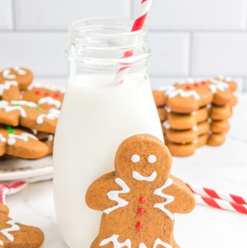 a gingerbread man cookie standing in front of a small bottle of milk with a red and white stray in it. In the background are stacks of decorated gingerbread men.