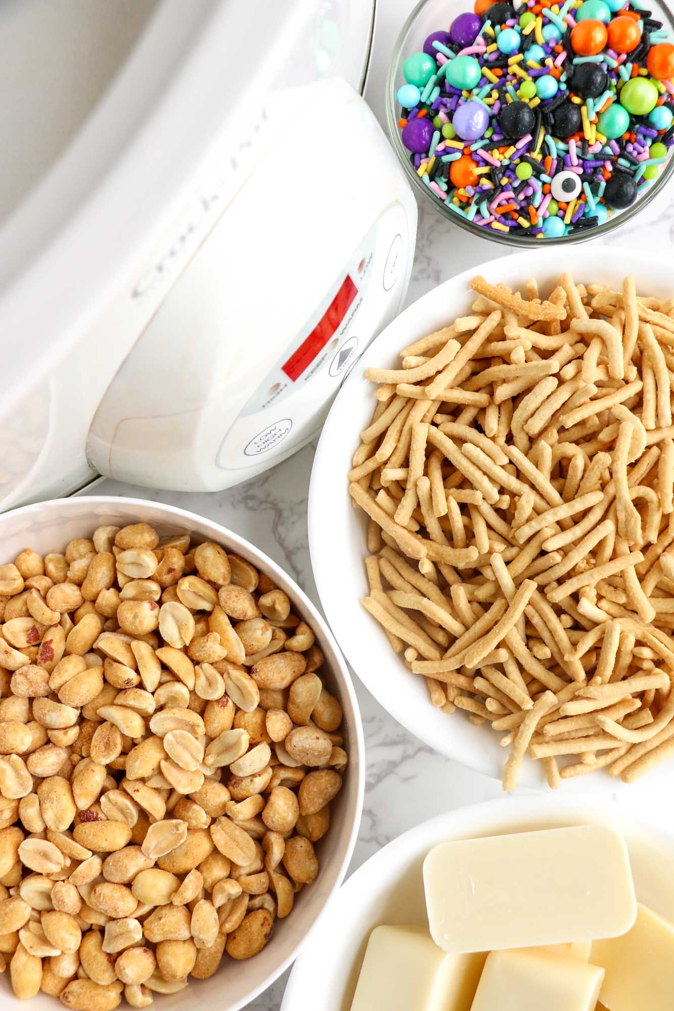 Ingredients needed to make halloween haystack cookies including chow mein noodles, roasted peanuts, white almond bark, and sprinkles.