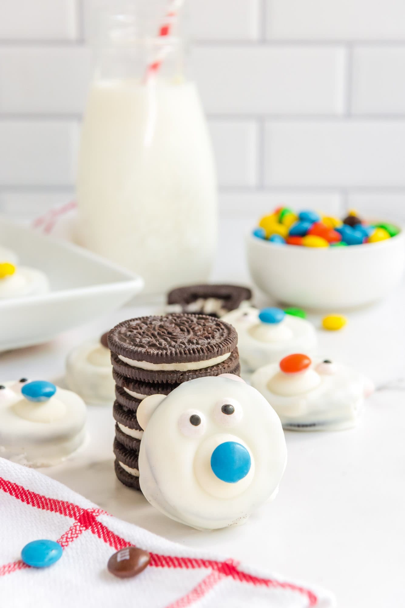 Polar bear cookie, and stacked oreo cookies in the background. And a bottle of milk with a straw in the background.
