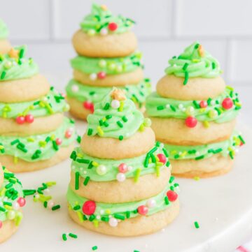 stacked christmas tree sugar cookies on a white tray. The cookies are filled with green frosting and decorated with holiday sprinkles.