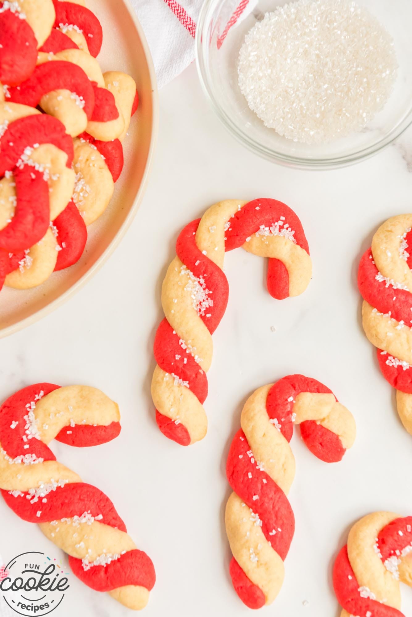 5 candy cane cookies on the counter next to a plate of the same. A bowl of coarse sugar is next to them.