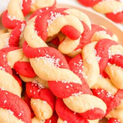Red and white swirled cookies shaped like candy canes, in a pile on a white plate.