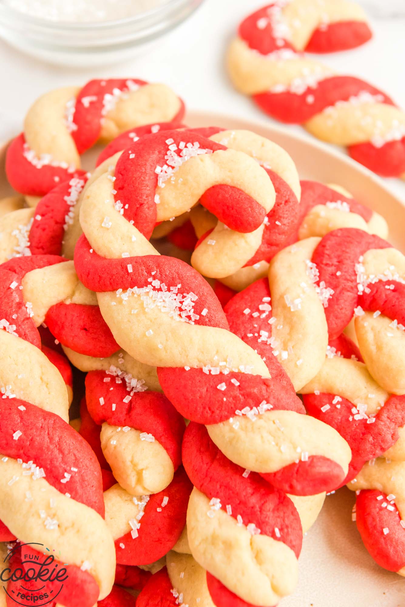 Red and white swirled cookies shaped like candy canes, in a pile on a white plate.