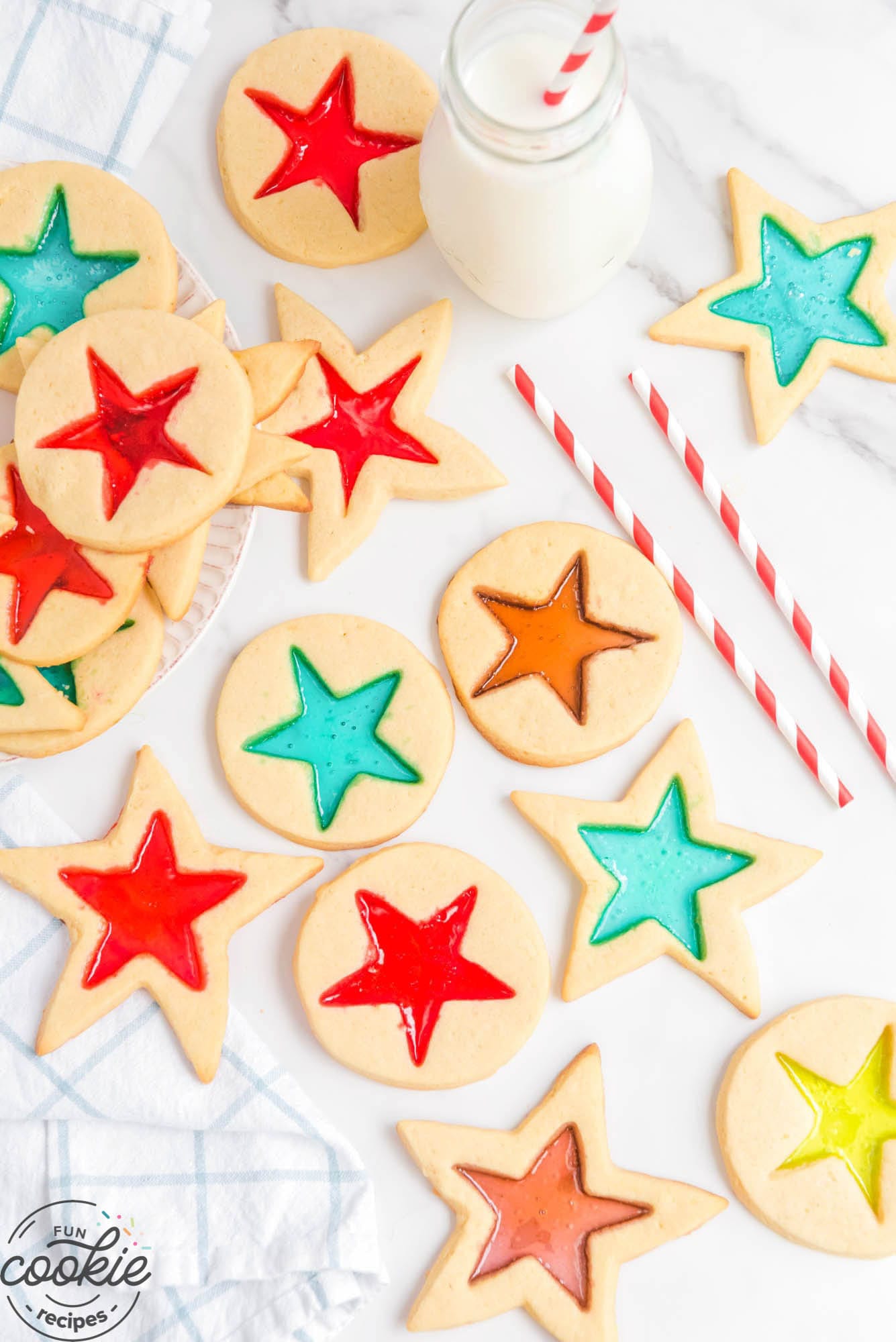 cookies cut out with stained glass stars on a counter next to a jar of milk and red and white striped straws.