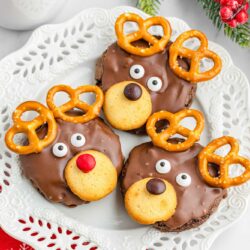 3 reindeer cookies on a white plate, with a mug of hot chocolate in the background