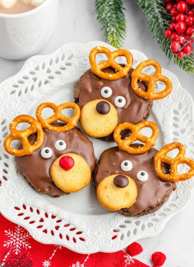 3 reindeer cookies on a white plate, with a mug of hot chocolate in the background