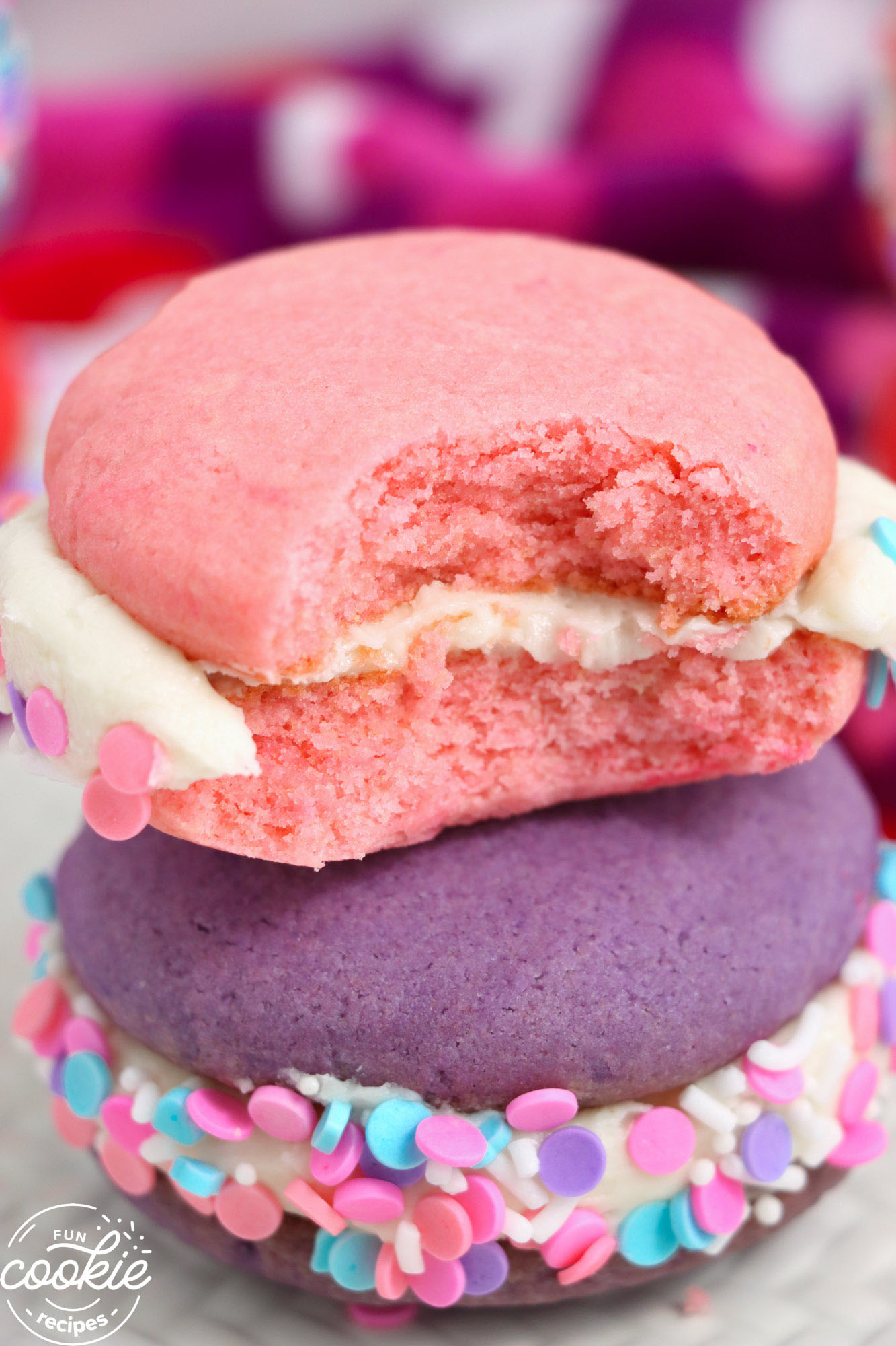 a pink whoopie pie on top of a p urple one. The cookies are filled with vanilla frosting and have sprinkles around the edges.