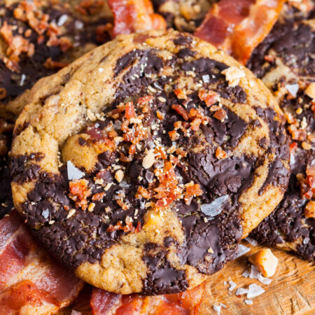 A large cookie with black and white dough, chocolate chunks and butterscotch chips, topped with salt and bacon, on top of strips of cooked bacon on a wooden board.
