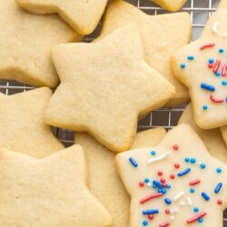 Mostly undecorated sugar cookies shaped like stars, placed on a wire rack. 2 of the cookies are glazed and decorated with blue and red sprinkles