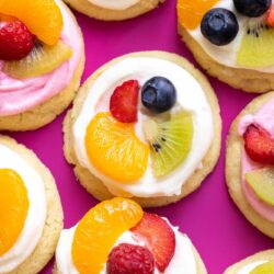 Fruit Pizza cookies on a dark pink background. Each cookie is topped with fluffy cream cheese frosting, mandarin oranges, and other fresh fruit pieces.