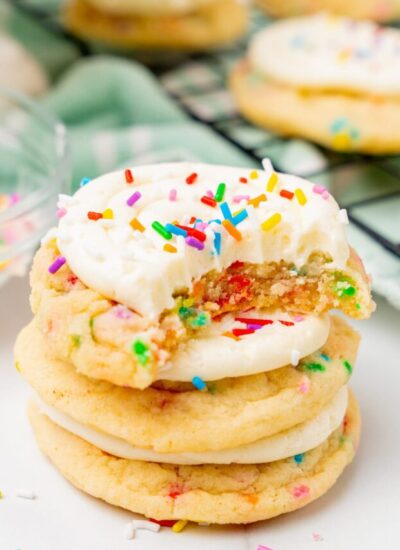 a stack of three frosted birthday cake cookies. The top one has been bitten into to show the sprinkles inside.