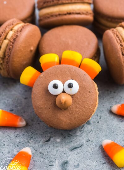 A brown macaron that is decorated to look like a thanksgiving turkey with candy eyes, and candy corn.