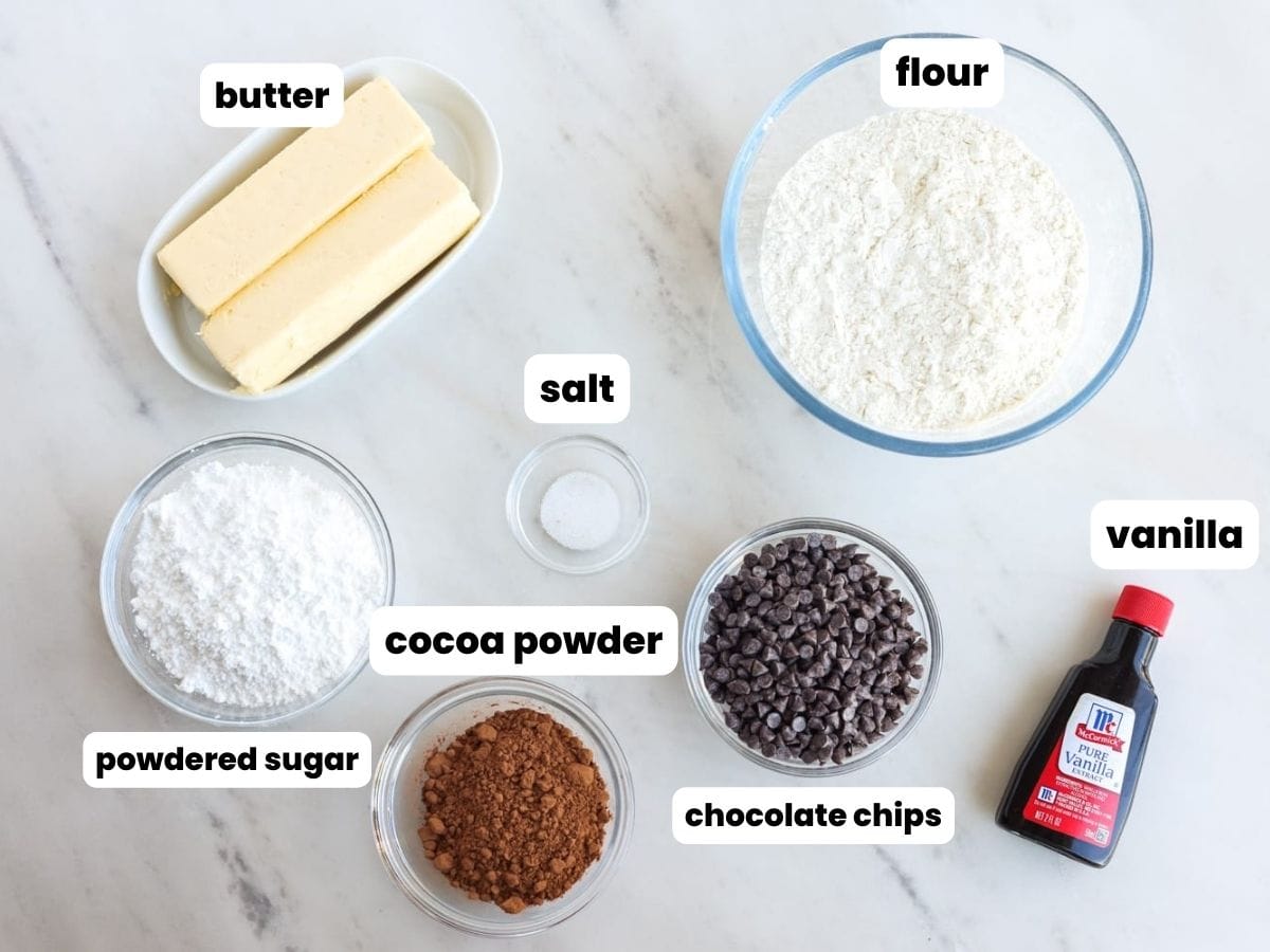 Ingredients needed to make chocolate snowball cookies, including butter, flour, powdered, sugar, cocoa powder, chocolate, chips, salt, and vanilla