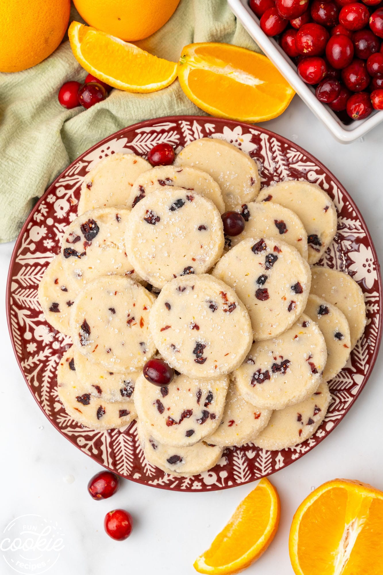 a red and white patterned plate holding a pile of round shortbread cookies with cranberries. Around the plate are fresh orange wedges and fresh cranberries.