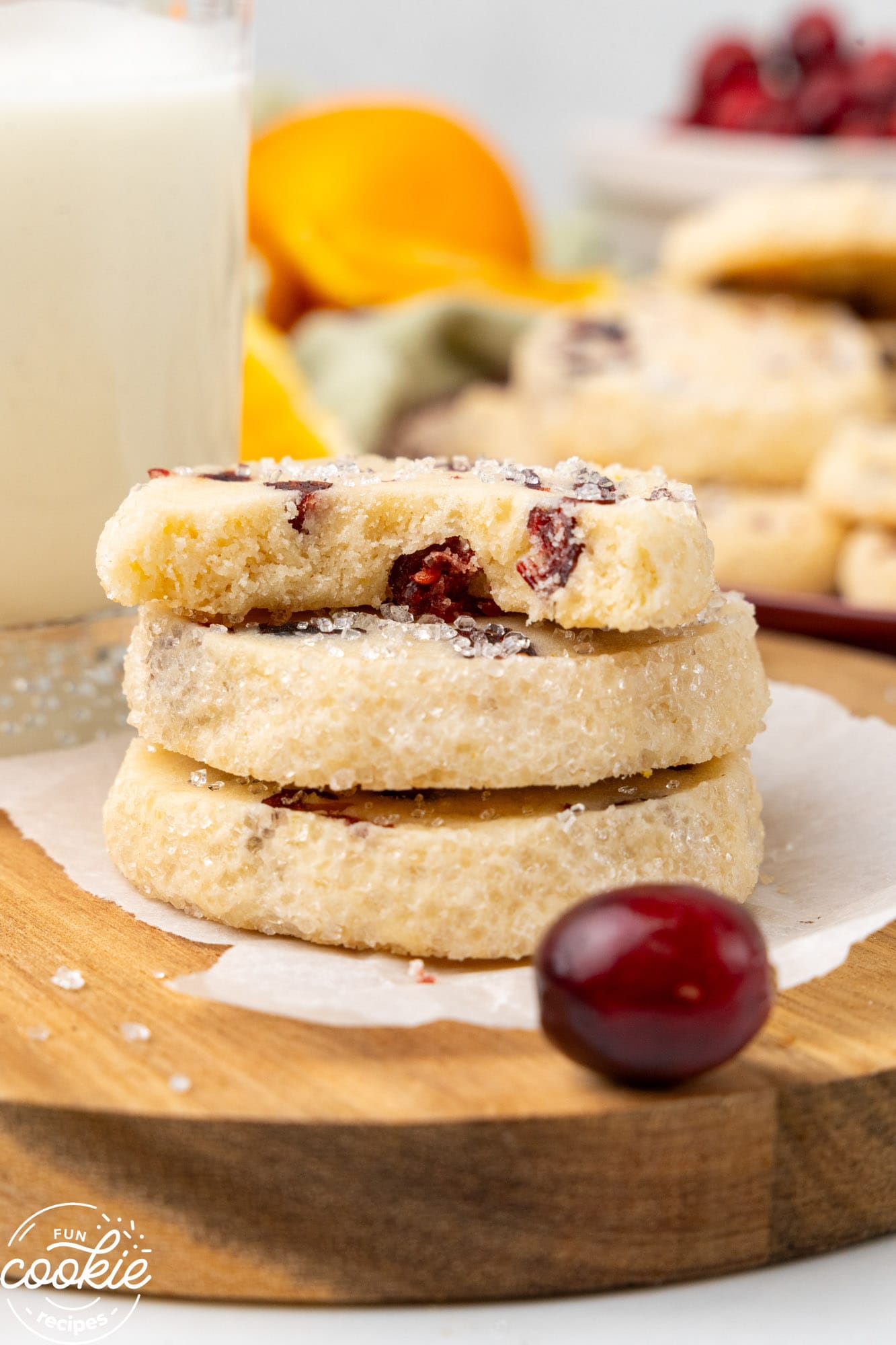 three cookies with cranberries and orange stacked. One has a bite taken. In the background is a glass of milk. 